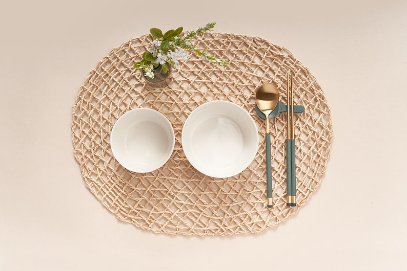 May include: Two white bowls, a gold spoon, and a pair of green chopsticks on a woven straw placemat. The bowls are empty and the spoon and chopsticks are resting on a small green holder.