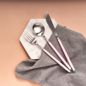 May include: A set of three pink-handled silverware pieces, including a fork, spoon, and knife, on a white marble hexagon-shaped plate. The silverware is arranged in a row, with the fork on the left, the spoon in the middle, and the knife on the right.