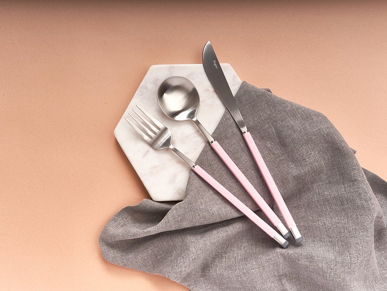 May include: A set of three pink-handled silverware pieces, including a fork, spoon, and knife, on a white marble hexagon-shaped plate. The silverware is arranged in a row, with the fork on the left, the spoon in the middle, and the knife on the right.
