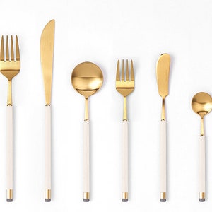 May include: A set of gold-toned flatware with white handles. The set includes a fork, knife, spoon, and chopsticks.