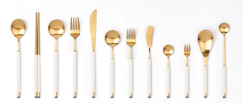 May include: A set of gold-toned flatware with white handles. The set includes a fork, knife, spoon, and chopsticks.
