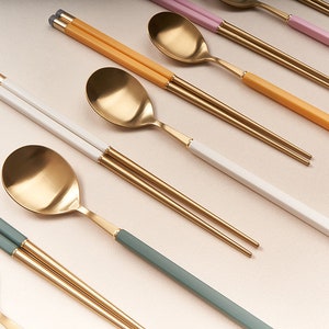 May include: A set of gold chopsticks and spoons with colorful handles. The handles are green, yellow, white, and pink. The chopsticks and spoons are arranged in a row on a light beige surface.
