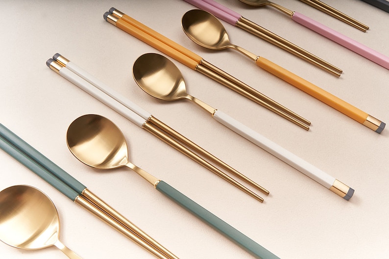 May include: A set of gold chopsticks and spoons with colorful handles. The handles are green, yellow, white, and pink. The chopsticks and spoons are arranged in a row on a light beige surface.