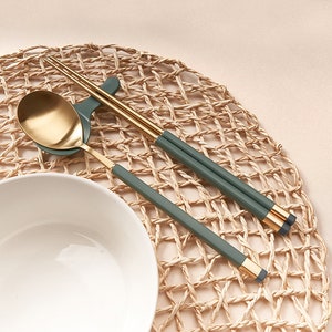 May include: A set of gold chopsticks and a gold spoon with green handles on a woven placemat. The chopsticks and spoon are resting on a white bowl.