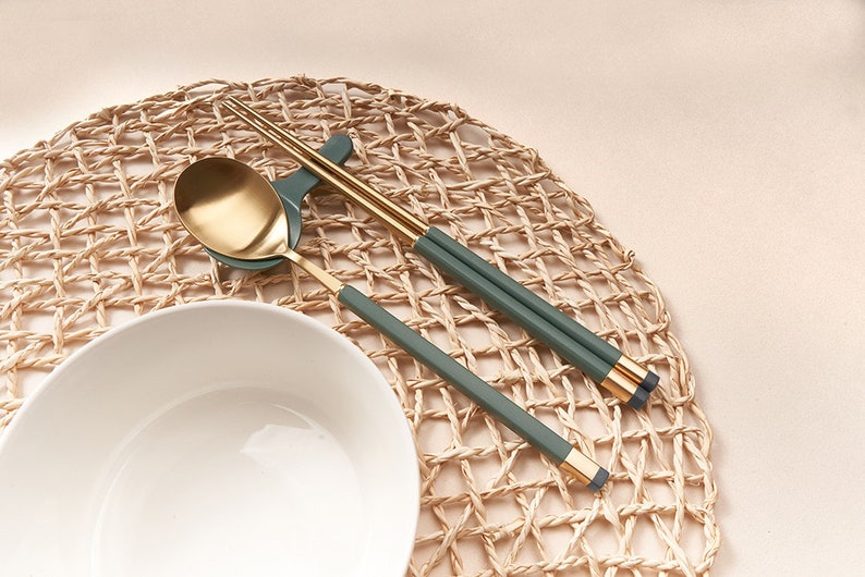 May include: A set of gold chopsticks and a gold spoon with green handles on a woven placemat. The chopsticks and spoon are resting on a white bowl.