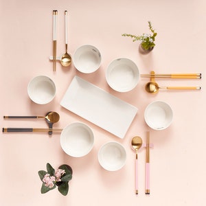 May include: A set of white bowls, gold chopsticks, and spoons arranged on a pink surface. The bowls are arranged in a circle with a white rectangular plate in the center. There are two small potted plants with pink flowers on the left and right sides of the image.