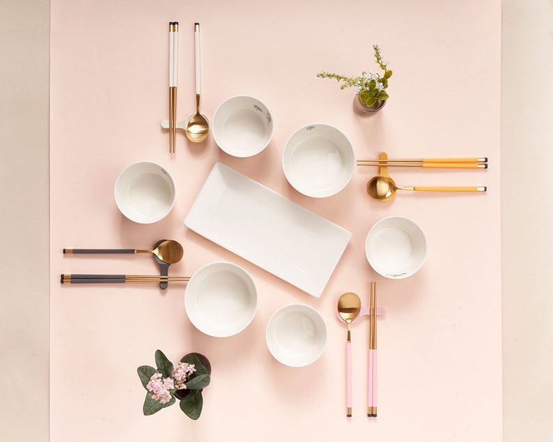 May include: A set of white bowls, gold chopsticks, and spoons arranged on a pink surface. The bowls are arranged in a circle with a white rectangular plate in the center. There are two small potted plants with pink flowers on the left and right sides of the image.
