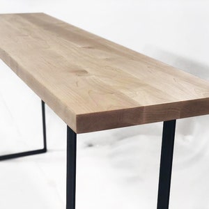 May include: A long, narrow table with a light wood top and black metal legs. The table is made of solid wood and has a simple, modern design.