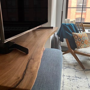 May include: A wooden TV stand with a live edge design. The stand is made of natural wood with a dark brown finish. The stand is supported by a black metal frame. The stand is in a living room setting with a sofa and a rug.