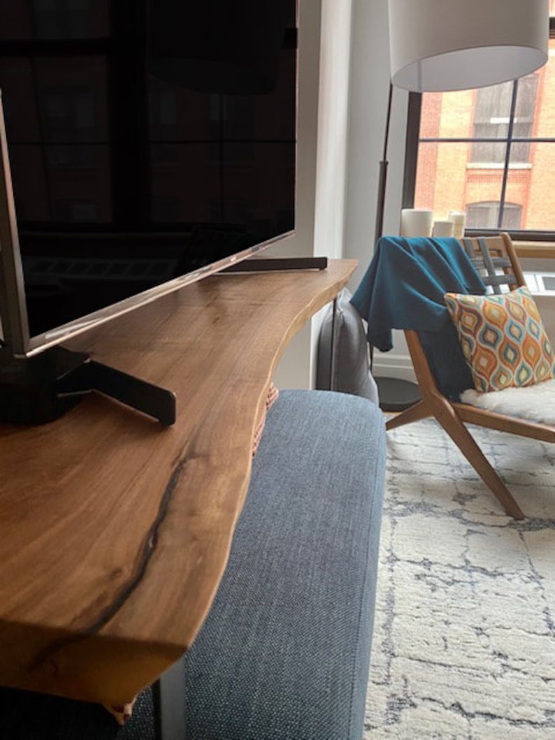 May include: A wooden TV stand with a live edge design. The stand is made of natural wood with a dark brown finish. The stand is supported by a black metal frame. The stand is in a living room setting with a sofa and a rug.