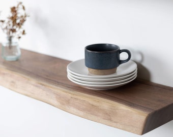 May include: A dark wood shelf with a dark blue ceramic mug and a stack of white ceramic saucers on top. The shelf is mounted on a white wall.