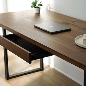 May include: A modern wooden desk with a dark brown finish and a black metal frame. The desk has a drawer that is partially open. A laptop, a small plant, a book, and a decorative knot are on the desk.