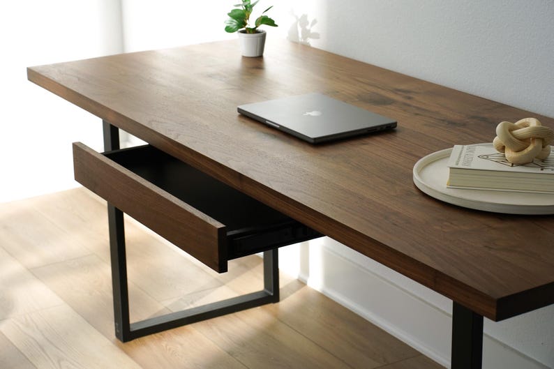 May include: A modern wooden desk with a dark brown finish and a black metal frame. The desk has a drawer that is partially open. A laptop, a small plant, a book, and a decorative knot are on the desk.