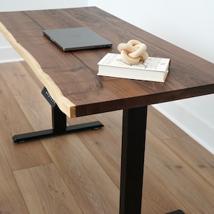 May include: A standing desk with a black metal frame and a natural wood top. The top has a live edge and is made of walnut wood. A laptop, a book, and a decorative knot are on the desk.