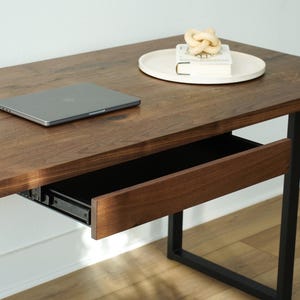 May include: A modern wooden desk with a pull-out drawer and black metal legs. A laptop sits on the desk surface, along with a decorative knot and a book on a white tray. The desk has a rich, warm wood tone.