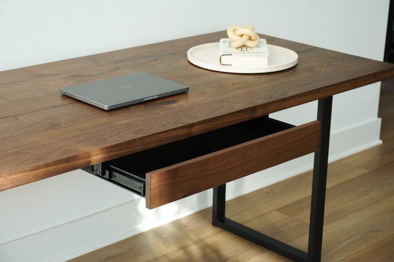 May include: A modern wooden desk with a pull-out drawer and black metal legs. A laptop sits on the desk surface, along with a decorative knot and a book on a white tray. The desk has a rich, warm wood tone.