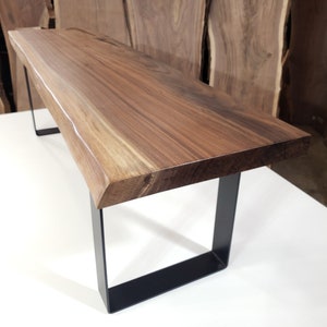 May include: A long, wooden bench with a live edge top and black metal legs. The bench is made of walnut wood and has a natural finish.