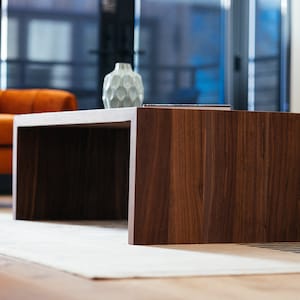 Pode incluir: Uma mesa de centro de madeira castanho escura com um tampo retangular e um espa&ccedil;o retangular aberto por baixo. A mesa est&aacute; sobre um tapete branco.