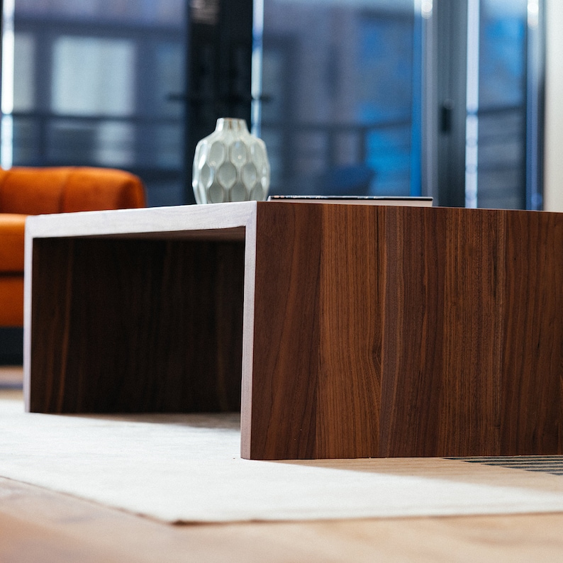Pode incluir: Uma mesa de centro de madeira castanho escura com um tampo retangular e um espa&ccedil;o retangular aberto por baixo. A mesa est&aacute; sobre um tapete branco.