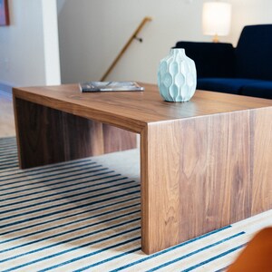 Pode incluir: Um vaso de cer&acirc;mica azul claro com uma superf&iacute;cie texturizada est&aacute; sobre uma mesa de centro de madeira marrom escura. A mesa tem uma superf&iacute;cie retangular larga e um design simples e moderno.