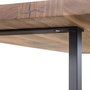 May include: Close-up of a wooden table with a natural wood grain finish. The table has a rectangular top and is supported by black metal legs. The legs are square and provide a modern, industrial aesthetic.