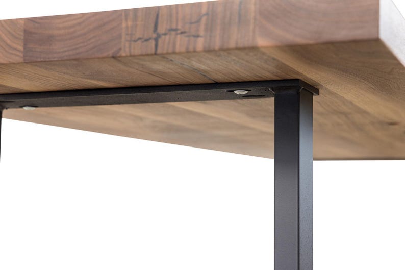 May include: Close-up of a wooden table with a natural wood grain finish. The table has a rectangular top and is supported by black metal legs. The legs are square and provide a modern, industrial aesthetic.
