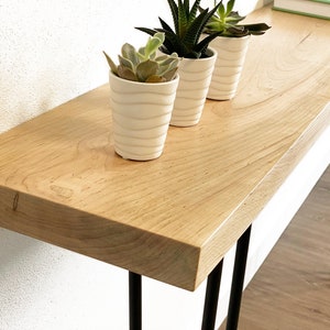 May include: A light wood console table with a natural finish and black hairpin legs. Three white ceramic planters with succulents are on the table.
