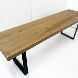 May include: A wooden bench with a natural wood finish and black metal legs. The bench is made of solid wood and has a simple, modern design.