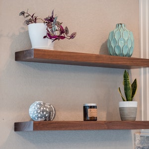 May include: Two wooden shelves with a white pot containing a trailing plant, a teal vase, a grey decorative sphere, and a candle on the lower shelf.