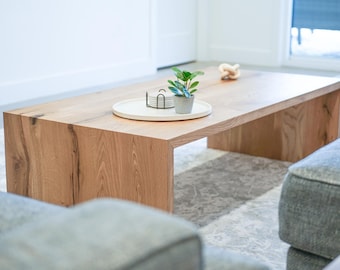 Puede incluir: Una mesa de centro de madera clara con una bandeja blanca y una pequeña planta en maceta en la parte superior. La mesa tiene una veta de madera natural y una forma rectangular. La mesa está sobre una alfombra gris claro.