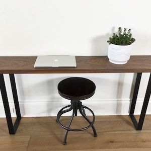 May include: A long, dark wood console table with black metal legs. A laptop computer is on the table, and a white planter with a green plant is on the right side of the table. A black metal stool is in front of the table.