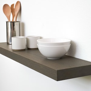 May include: A dark grey floating shelf with a wood grain texture. The shelf is mounted to a white wall and has a variety of kitchen items on it, including wooden spoons, white ceramic mugs, and white ceramic bowls.