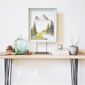 May include: A wooden console table with black hairpin legs. The table is decorated with a framed painting of a mountain landscape, a glass vase with a beaded garland, pine cones, and a small potted plant.