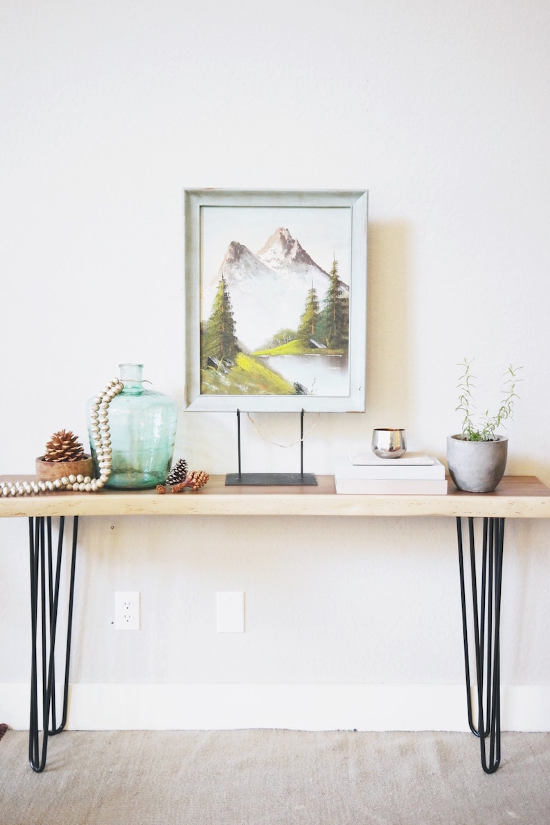 May include: A wooden console table with black hairpin legs. The table is decorated with a framed painting of a mountain landscape, a glass vase with a beaded garland, pine cones, and a small potted plant.