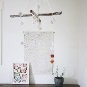 May include: A white wooden branch with white pom poms hanging from it. The branch is hanging from the ceiling and is centred in the image. There is a white canvas with a light grey pattern hanging on the wall behind the branch. A framed print of colourful butterflies is on a wooden table below the canvas. A small potted plant is on the table to the right of the print.