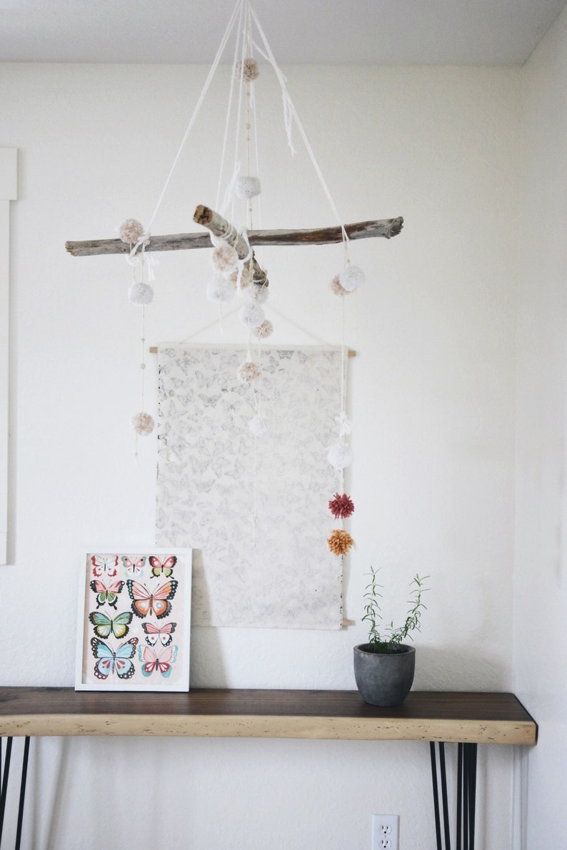 May include: A white wooden branch with white pom poms hanging from it. The branch is hanging from the ceiling and is centred in the image. There is a white canvas with a light grey pattern hanging on the wall behind the branch. A framed print of colourful butterflies is on a wooden table below the canvas. A small potted plant is on the table to the right of the print.