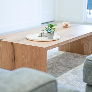 Pode incluir: Uma mesa de centro de madeira clara com uma bandeja branca e uma pequena planta em vaso no topo. A mesa tem um gr&atilde;o de madeira natural e uma forma retangular. A mesa est&aacute; sobre um tapete cinza claro.