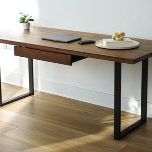 May include: A modern wooden desk with a dark brown finish and black metal legs. The desk has a built-in drawer, a laptop, a remote, a decorative object, and a small plant. The desk is approximately 60 inches long.