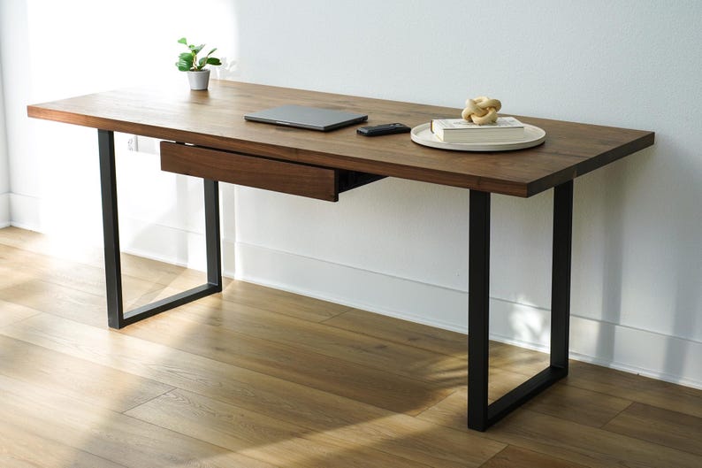 May include: A modern wooden desk with a dark brown finish and black metal legs. The desk has a built-in drawer, a laptop, a remote, a decorative object, and a small plant. The desk is approximately 60 inches long.