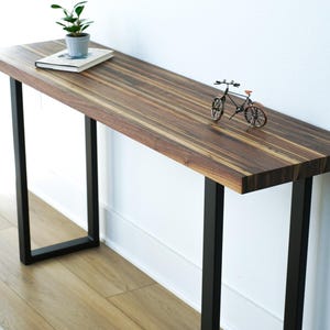 May include: A modern console table with a dark wood grain top and black metal legs. A small potted plant, a book, and a miniature bicycle are displayed on the table. The table is approximately 48 inches long.