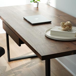 May include: A dark brown wooden desk with a black metal frame. A silver laptop, a white tray with a book and a decorative knot are on the desk. A black chair with gold accents is in the foreground.