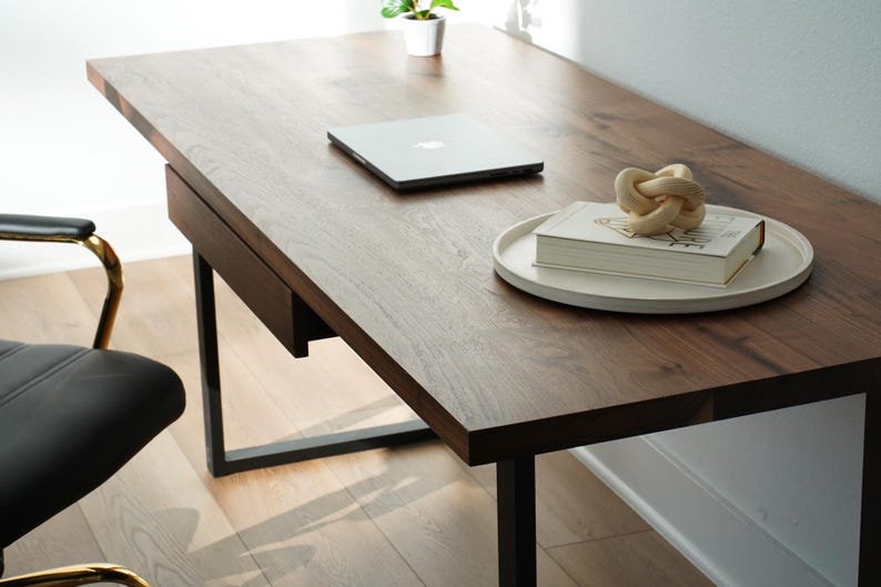 May include: A dark brown wooden desk with a black metal frame. A silver laptop, a white tray with a book and a decorative knot are on the desk. A black chair with gold accents is in the foreground.