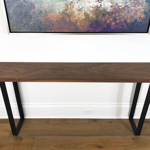 May include: A long, dark brown wooden console table with black metal legs. The table is sitting on a hardwood floor.