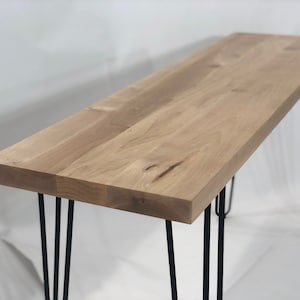 May include: A long, rectangular wooden table with a natural wood finish. The table has four black hairpin legs.