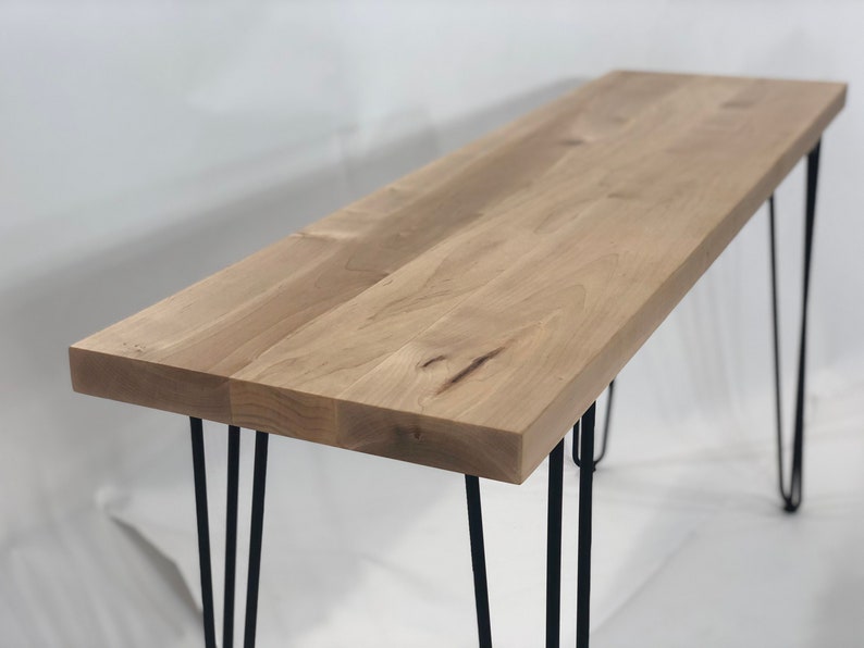 May include: A long, rectangular wooden table with a natural wood finish. The table has four black hairpin legs.
