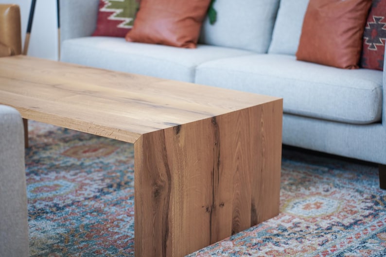 Pode incluir: Uma mesa de centro de madeira castanho claro com um tampo retangular e um design &uacute;nico de lados abertos. A mesa tem um padr&atilde;o de gr&atilde;o de madeira natural e est&aacute; sobre um tapete colorido.