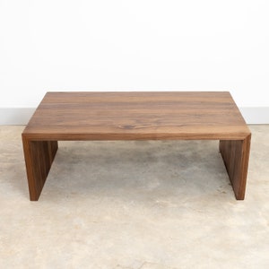Pode incluir: Uma mesa de centro de madeira castanho escura com tampo retangular e um design de moldura aberta &uacute;nico. A mesa tem um acabamento liso e polido e est&aacute; sobre um piso de concreto cinza claro.