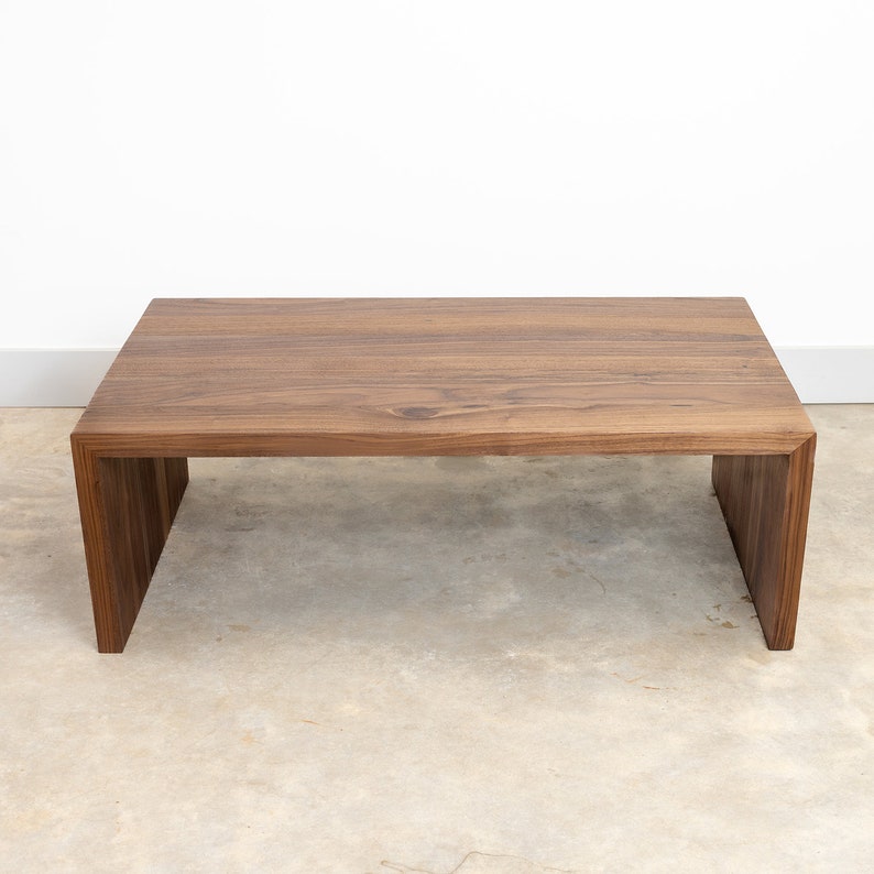 Pode incluir: Uma mesa de centro de madeira castanho escura com tampo retangular e um design de moldura aberta &uacute;nico. A mesa tem um acabamento liso e polido e est&aacute; sobre um piso de concreto cinza claro.