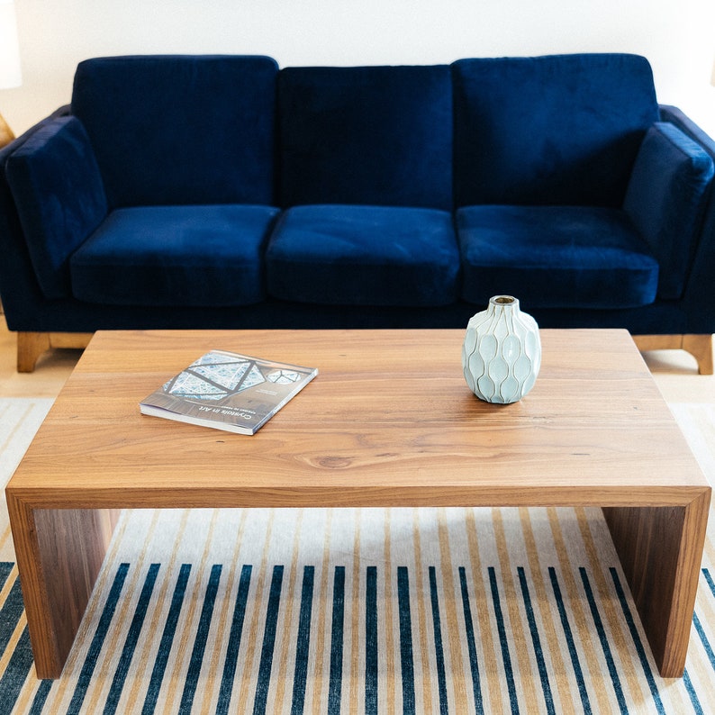 Pode incluir: Uma mesa de centro de madeira com acabamento em madeira clara e um vaso de cer&acirc;mica branco com um design texturizado. Um livro est&aacute; colocado sobre a mesa. A mesa est&aacute; na frente de um sof&aacute; de veludo azul.