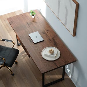May include: An overhead shot of a home office setup. A dark wood desk with black metal legs holds a laptop, a small plant, and a plate with a decorative object. A black leather office chair with gold accents sits to the left.